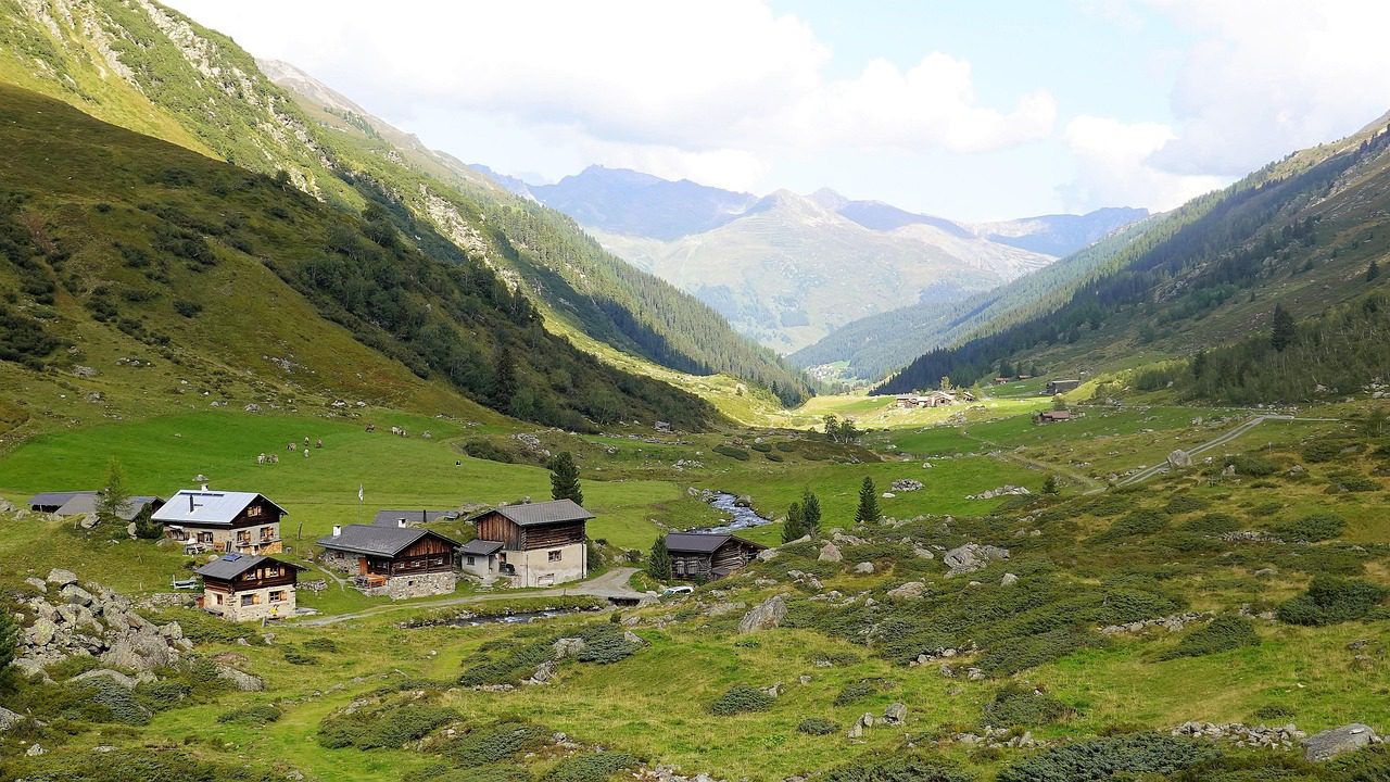 villiage in a valley in possibly the alps. a few houses nestled in a valley with a pictureqses series of mountains and clouds/fog in the background